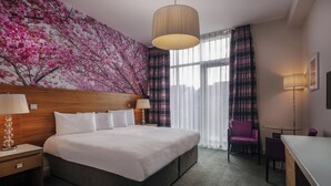 Una habitación de hotel con una cama grande, un cabecero de madera, un mural floral rosa en la pared, un ventanal con cortinas y un escritorio con una silla.