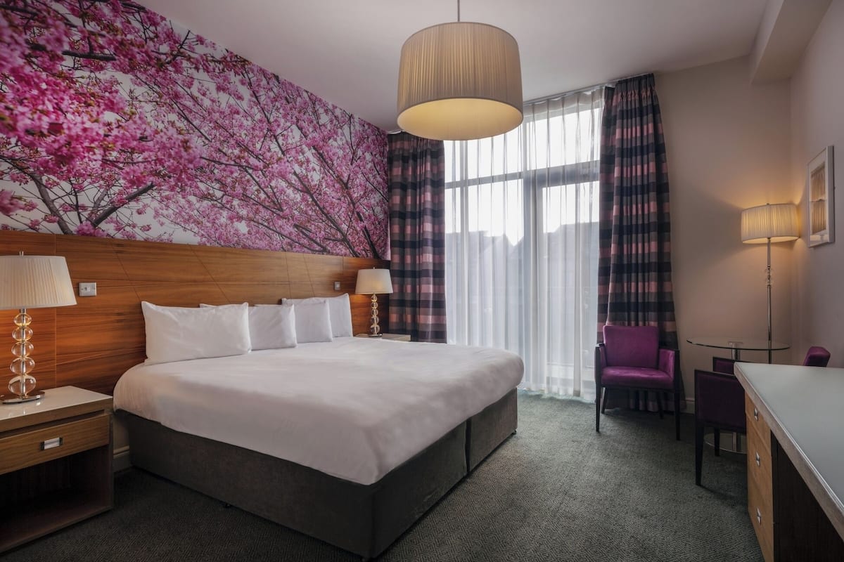 A hotel room with a large bed, a wooden headboard, a wall mural of pink flowers, a large window with curtains, and a desk with a chair.
