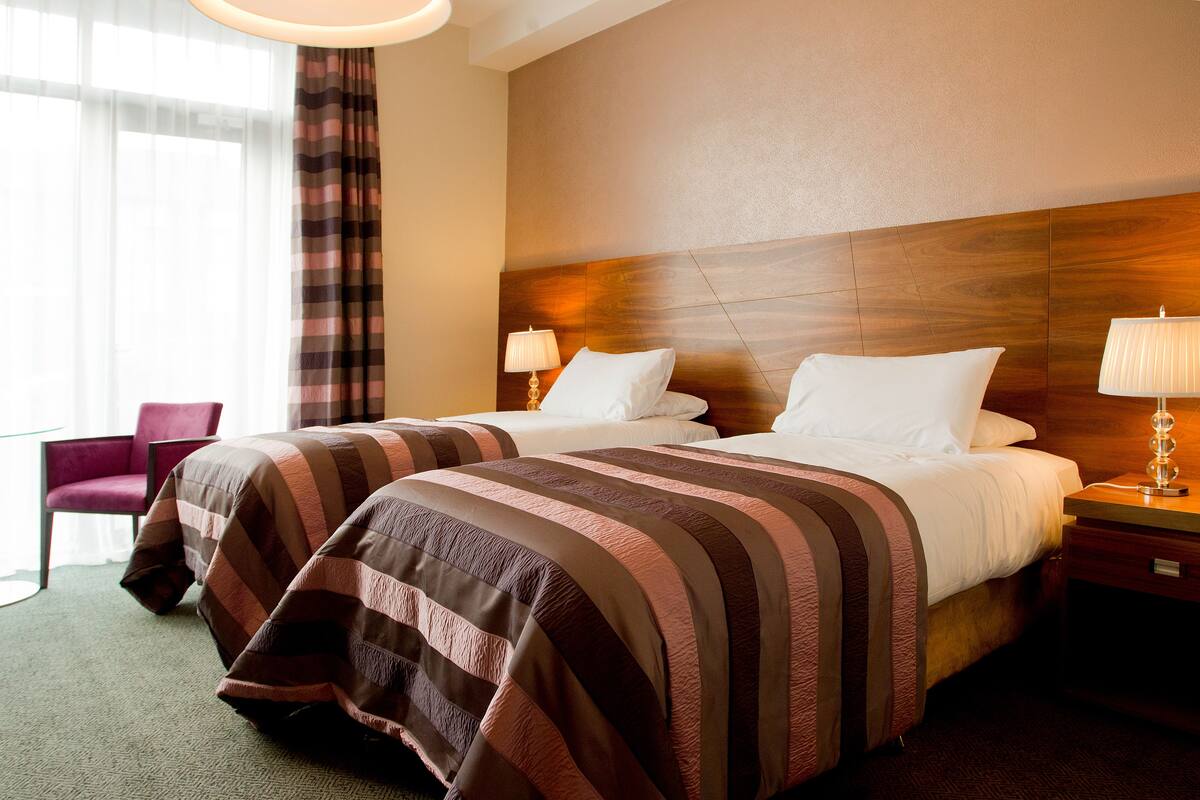 A hotel room with two beds, a wooden headboard, a chair, and a nightstand with a lamp.
