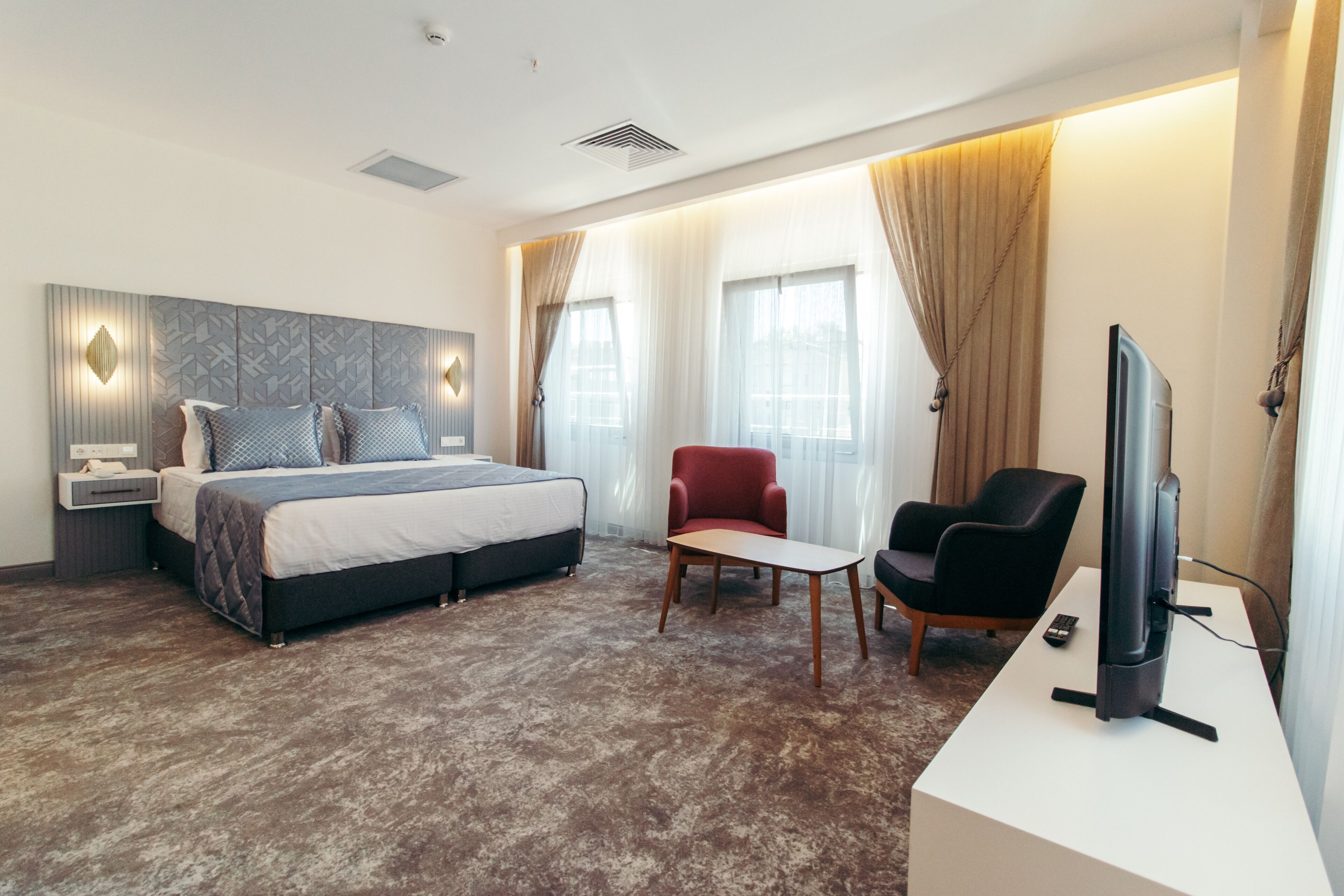 A modern hotel room with a large bed, a TV, a red armchair, and a small table.