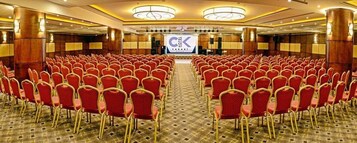 Un amplio salón de conferencias con filas de sillas rojas, un escenario con un logotipo y una alfombra estampada.