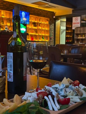 A bottle of wine, a glass, and a cheese board with various cheeses and fruits on a table in a restaurant.