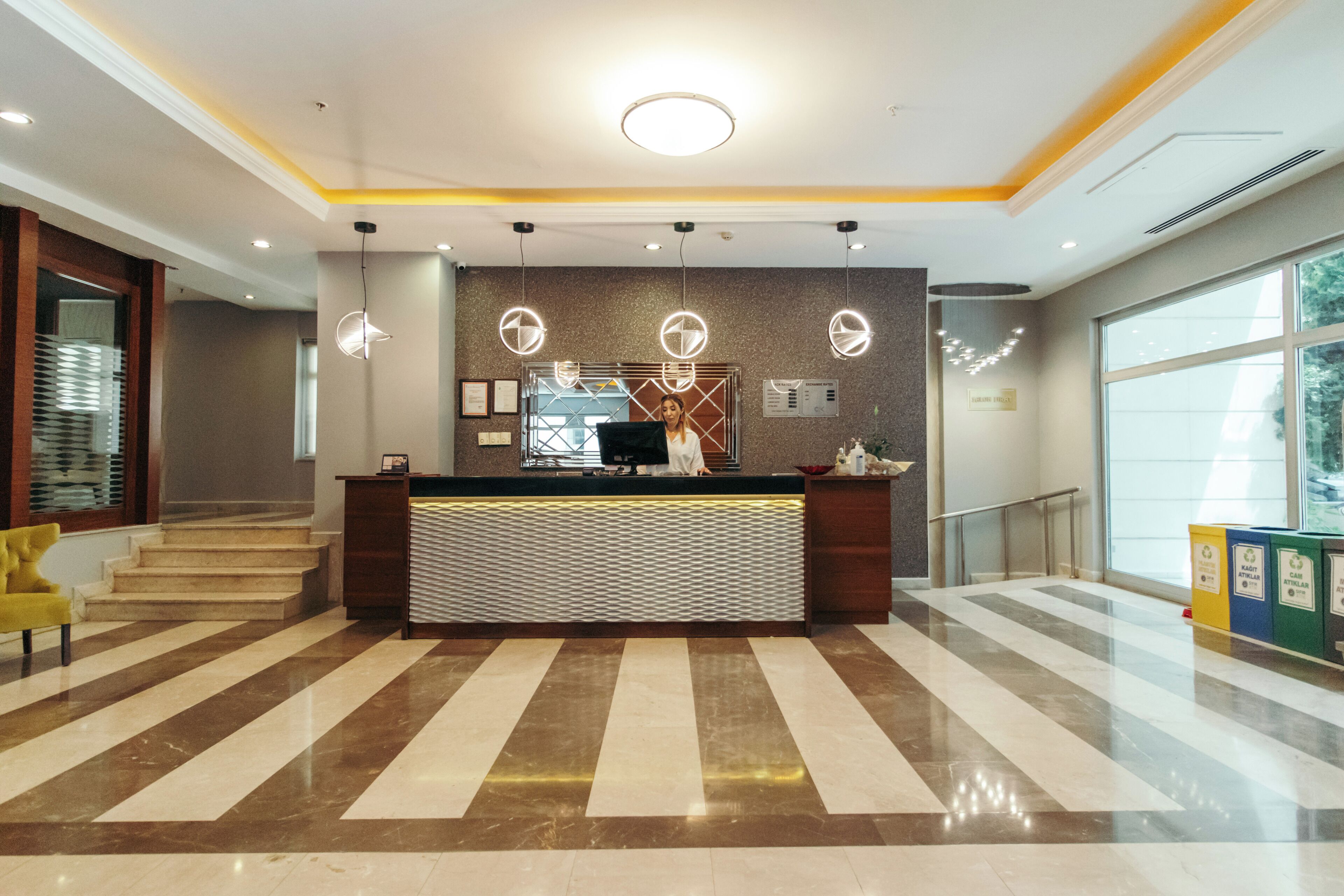 A hotel reception area with a modern design, featuring a reception desk, a staircase, and a large window providing natural light.