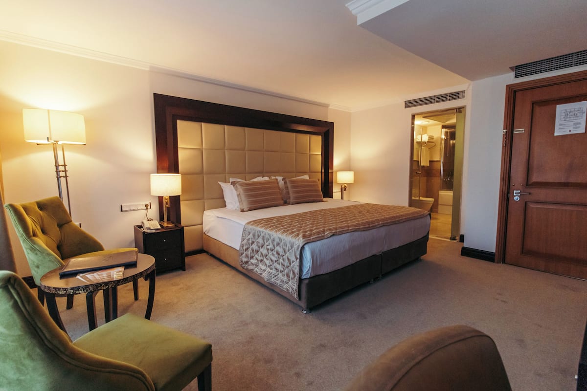 A hotel room with a large bed, two bedside lamps, a desk with a laptop, and a green armchair.