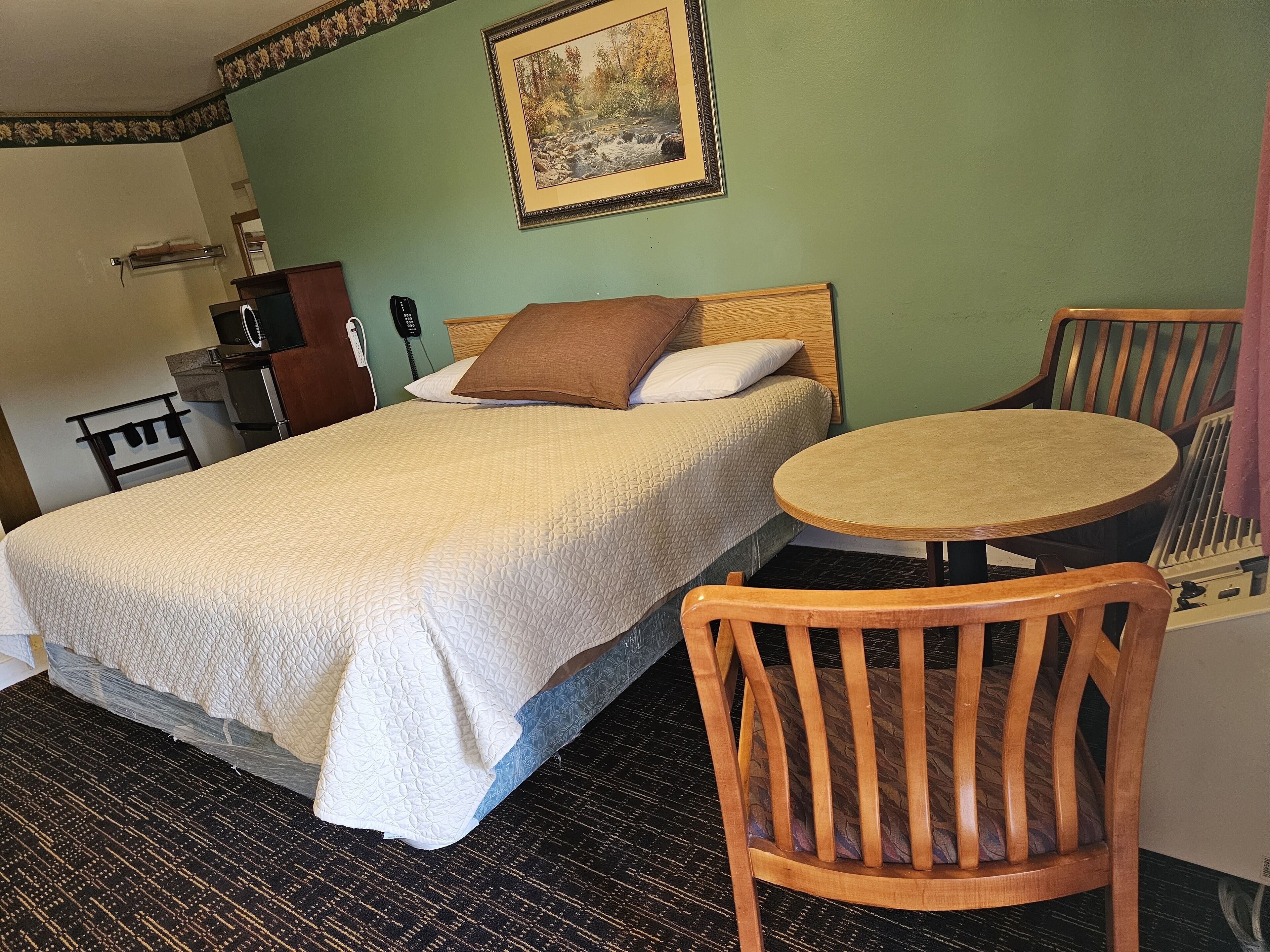 A hotel room with a bed, a round wooden table, a wooden chair, a desk with a telephone, and a framed picture on the wall.