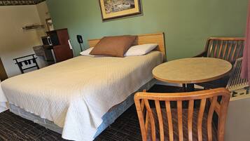 A hotel room with a bed, a round wooden table, a wooden chair, a desk with a telephone, and a framed picture on the wall.