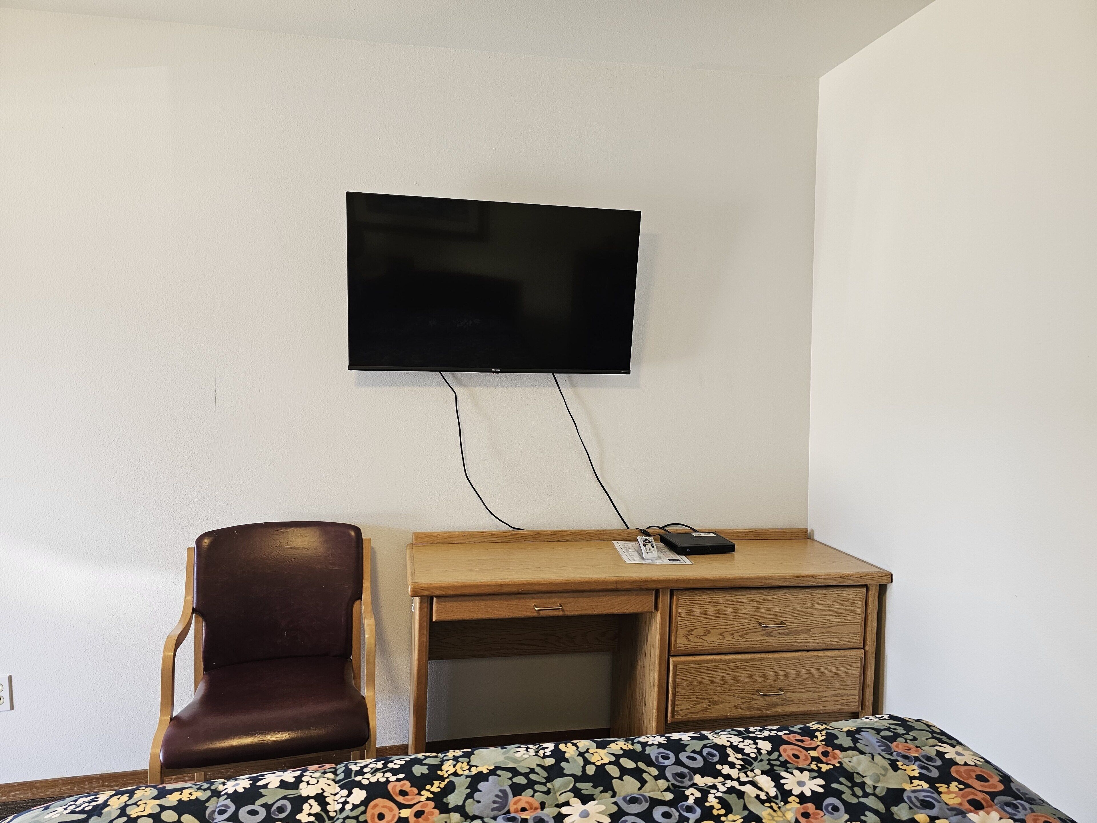 A hotel room with a wooden desk, a leather chair, a bed with a floral bedspread, and a mounted television.