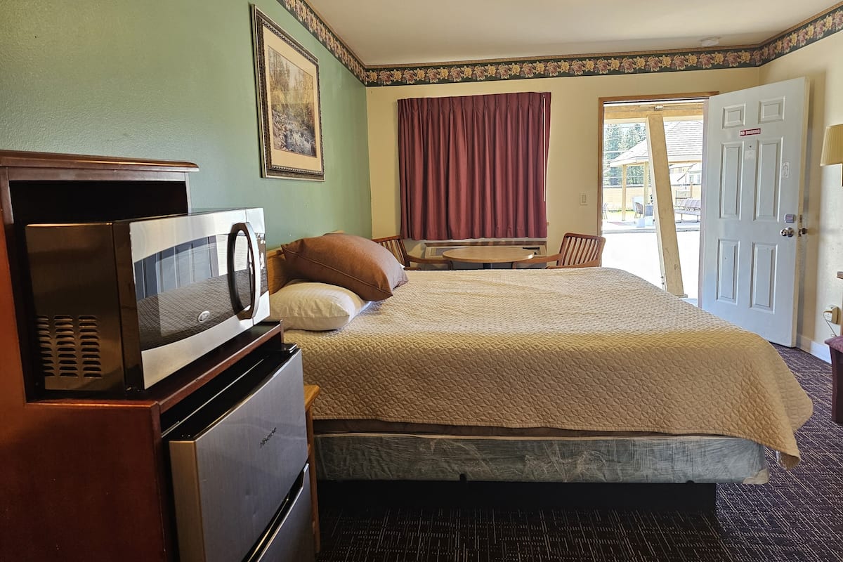 A hotel room with a bed, a desk with a microwave, a chair, a window with curtains, and a door with a 'No Smoking' sign.