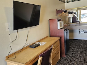 A hotel room with a wooden desk, a television mounted on the wall, a microwave, a refrigerator, and a sink with a mirror above it.