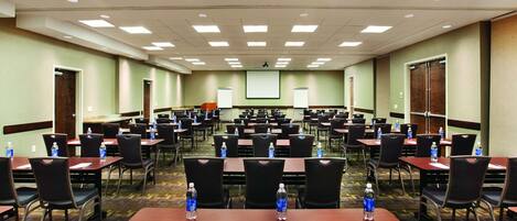 A conference room with multiple tables and chairs, equipped with water bottles and papers, and a projector screen at the front.