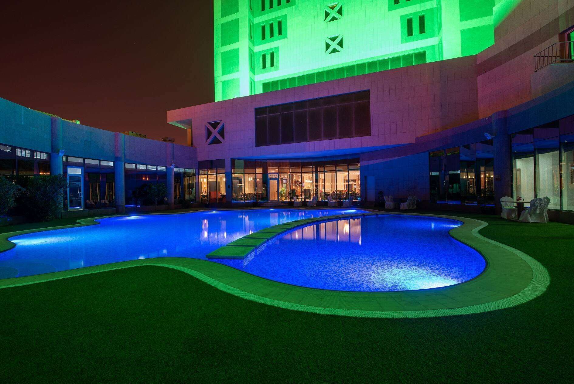 A nighttime view of a hotel with a swimming pool, a covered outdoor area, and a building with illuminated windows.