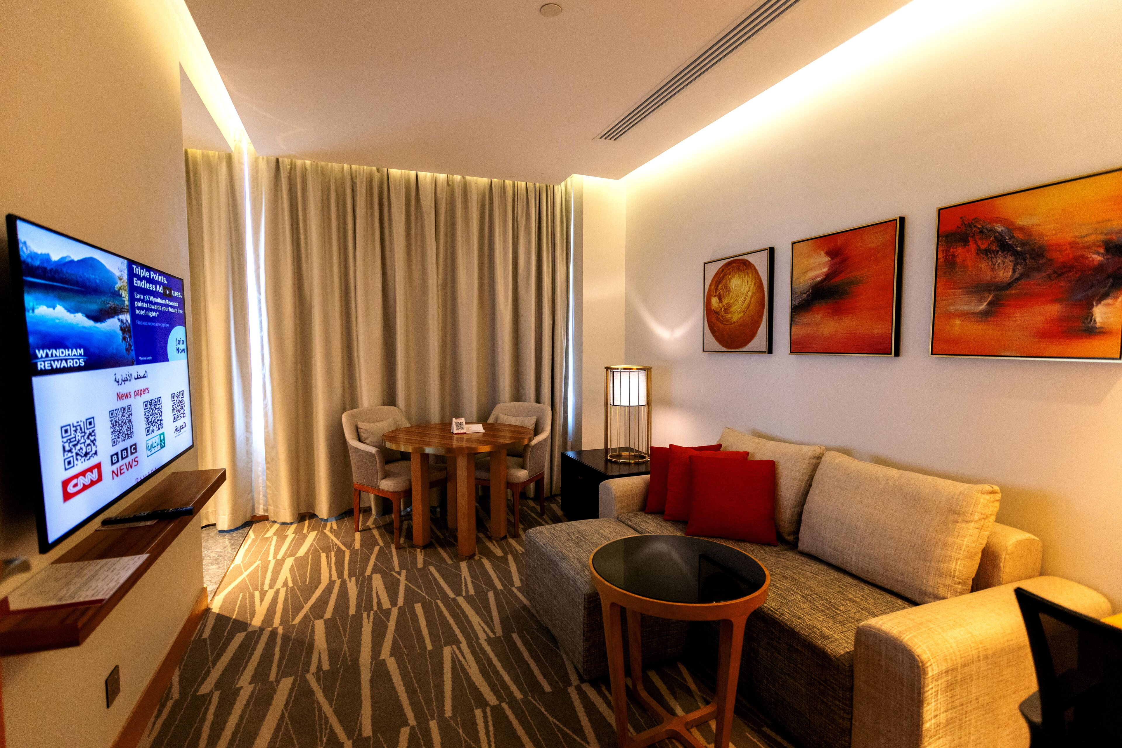 Habitación de hotel con televisión, una mesa de comedor con sillas, un sofá y tres cuadros en la pared.