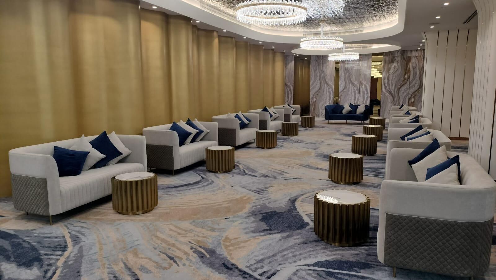 A hotel lobby with sofas and round tables arranged in a row, a marble-patterned carpet, and a chandelier.
