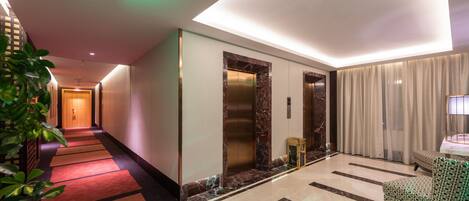 A hotel lobby with a long corridor, elevators, a reception area, and a seating area.