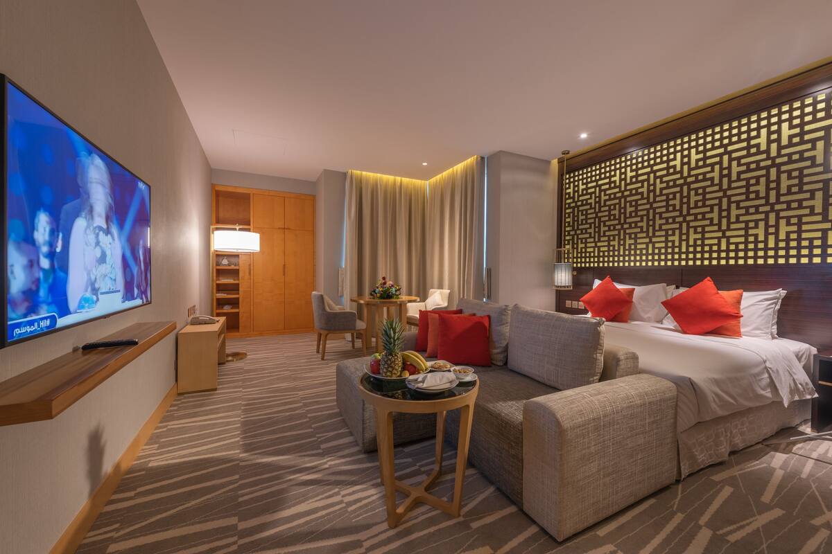 Ein Hotelzimmer mit Fernseher, einer Couch, einem Bett mit roten Kissen, einem hölzernen Kopfteil, einer Wand mit Muster und einem kleinen Tisch mit Früchten und Tablett.