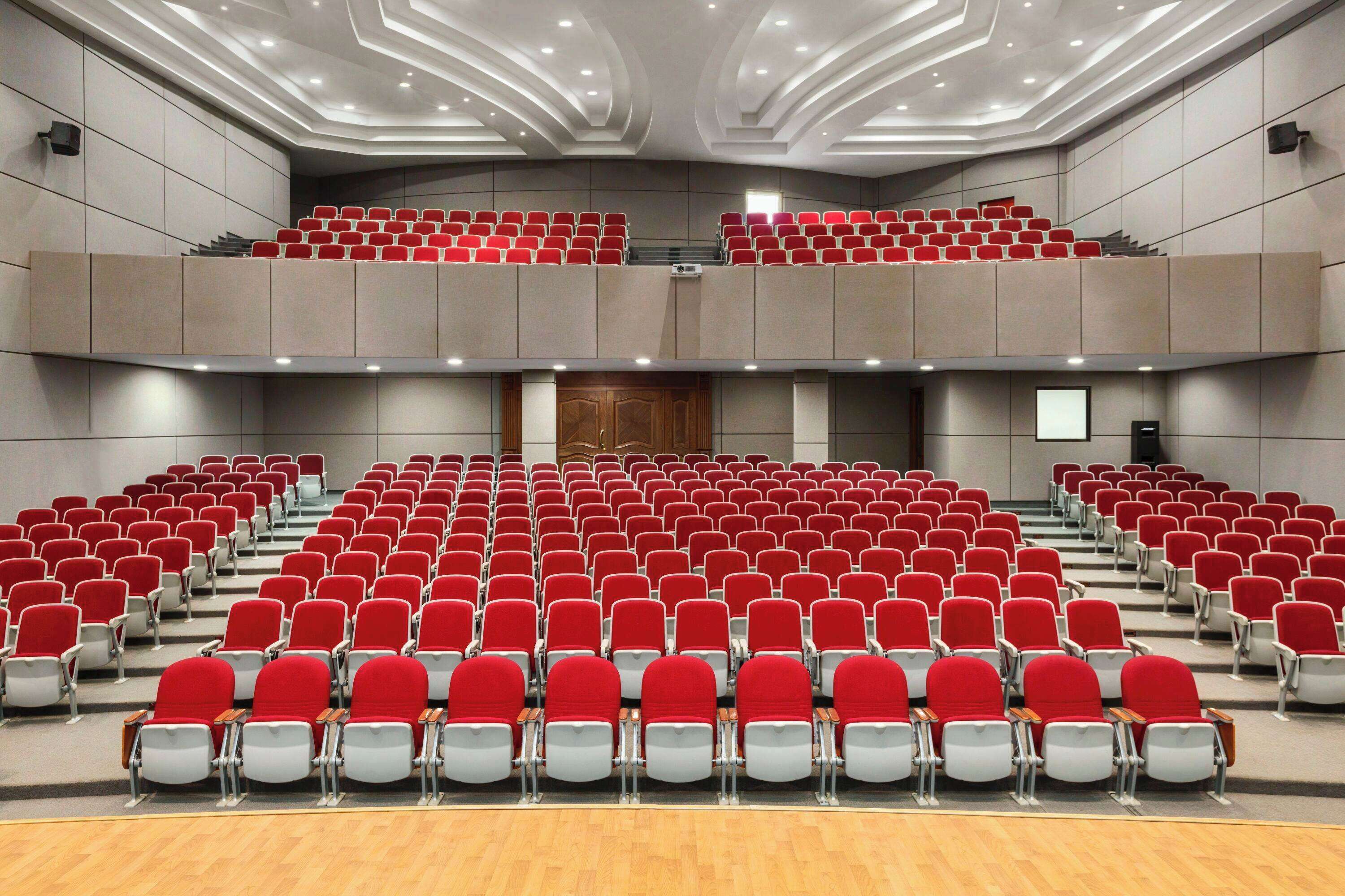 A large auditorium with rows of red and grey chairs, a wooden floor, and a modern ceiling design.