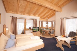 A cozy room with a wooden ceiling, a sofa with pillows, a television, a dining table with chairs, and a window with curtains. - Hotel Landhaus Feldmeier (Oberammergau)