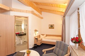 A compact hotel room with a single bed, a small bedside table, a chair, a window with curtains, and a view of the outdoors. - Hotel Landhaus Feldmeier (Oberammergau)