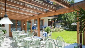 Un patio al aire libre con mesas y sillas, una pérgola de madera y vistas a un jardín.