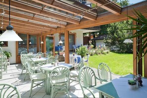An outdoor patio with tables and chairs, a wooden pergola, and a view of a garden. - Hotel Landhaus Feldmeier (Oberammergau)