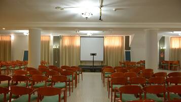 Un salón de conferencias con hileras de sillas mirando hacia una pantalla de proyección, un ventilador de techo y una zona de escenario.