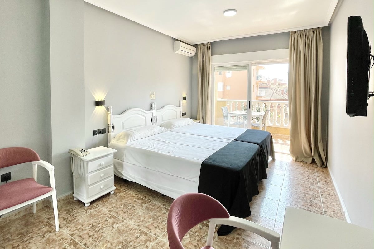 A hotel room with a large bed, a desk, a chair, a bedside table, and a balcony with a table and chairs.