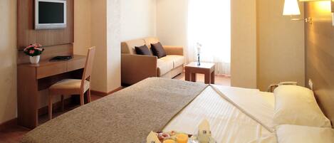 A hotel room with a bed, a desk, a chair, a television, a small table with a vase, and a tray with breakfast items.