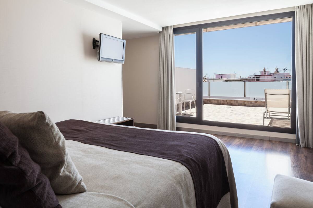 Habitación de hotel con cama, televisor en la pared, balcón con mesa y sillas, y vista al mar.