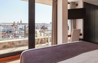 Une chambre d’hôtel avec un grand lit, une vue sur le paysage urbain, un balcon avec des chaises et une armoire sombre.