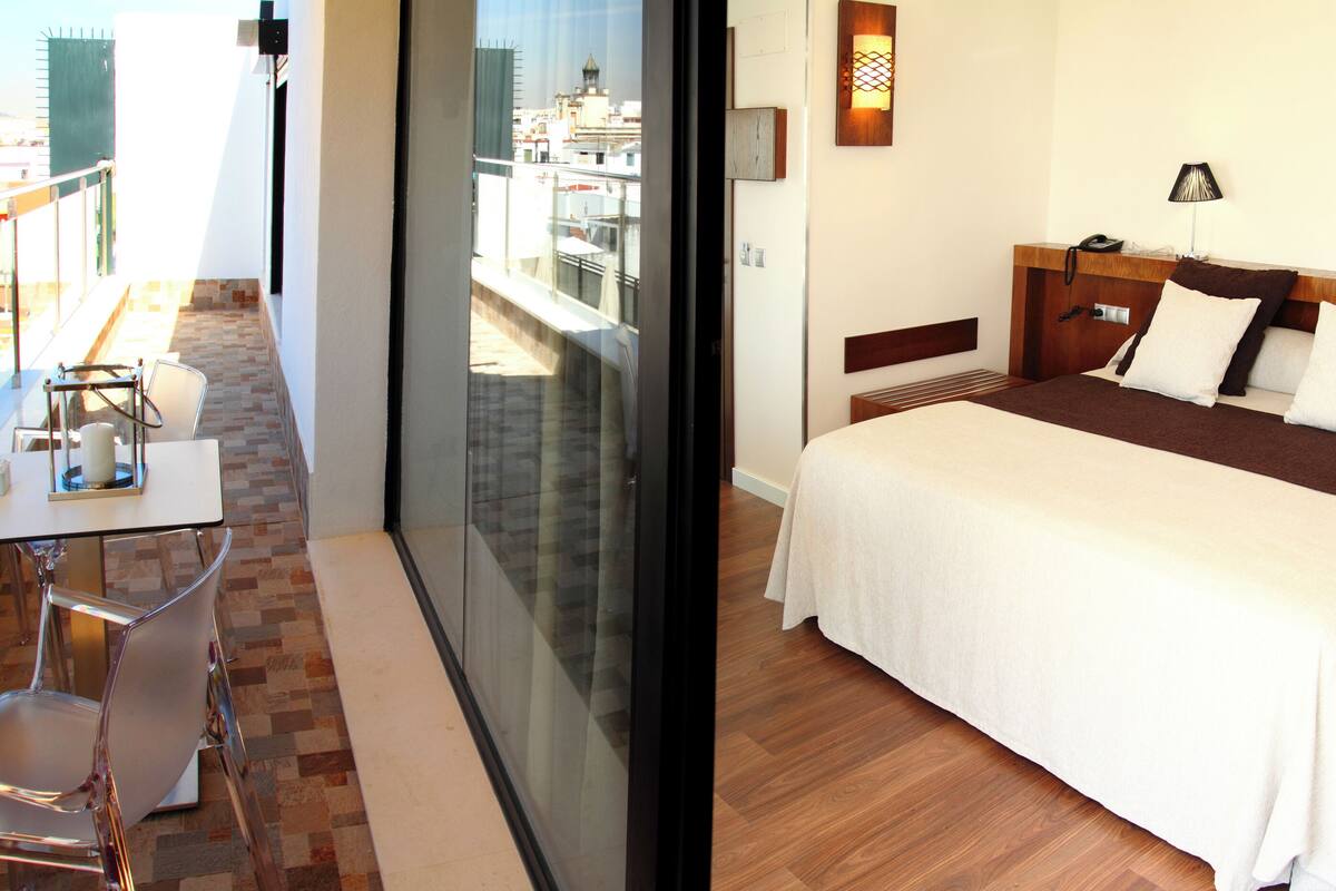Una habitación de hotel con balcón, una cama con muchos almohadones, una mesita de noche y una lámpara.