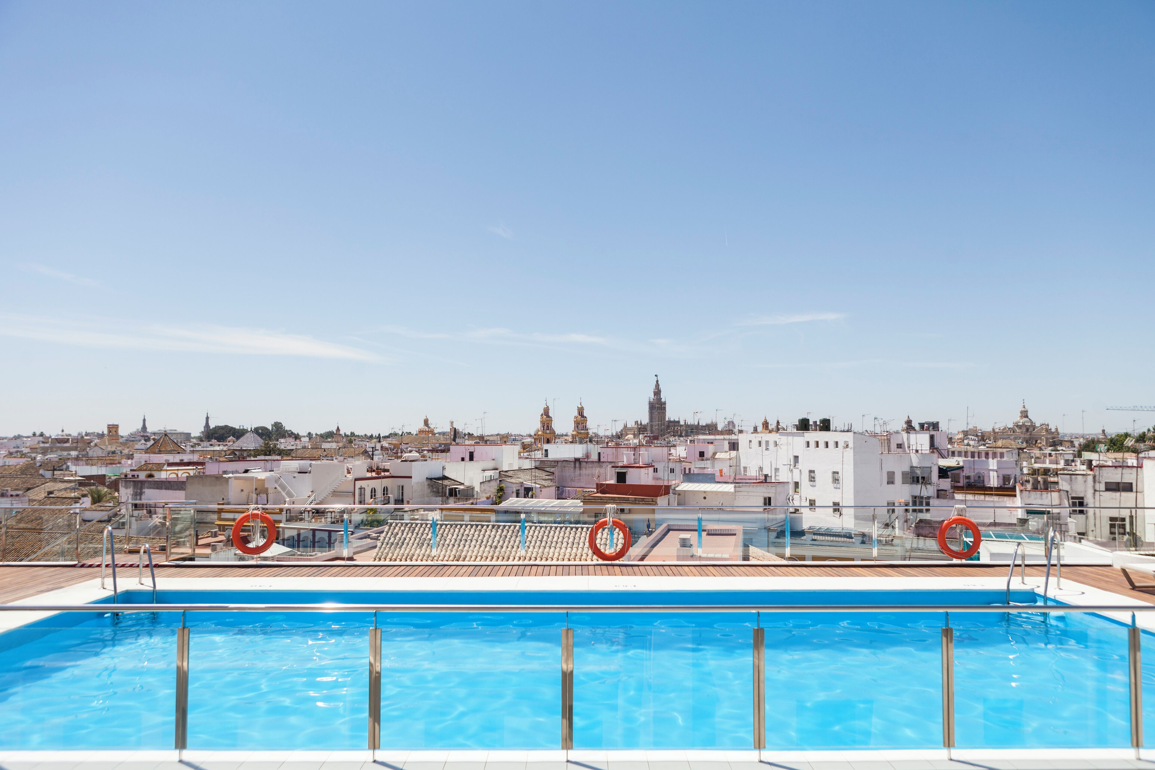 A rooftop swimming pool with clear blue water, surrounded by a metal railing. In the background, there is a view of a cityscape with various buildings and a prominent tower.