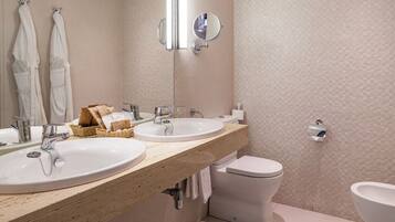 A modern bathroom with two sinks, a toilet, and a bidet. There are towels hanging on hooks and a basket with toiletries on the countertop.