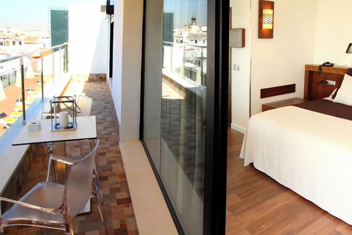 A balcony with a table and chairs, a bed with a wooden headboard, and a city view.