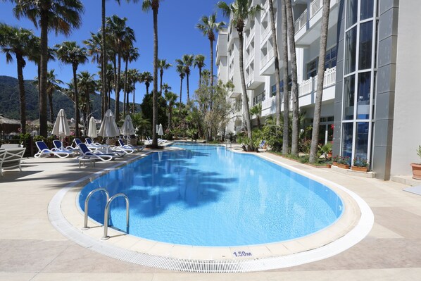 Une piscine entourée de palmiers et d’un bâtiment d’hôtel, avec des chaises longues et des parasols à proximité.