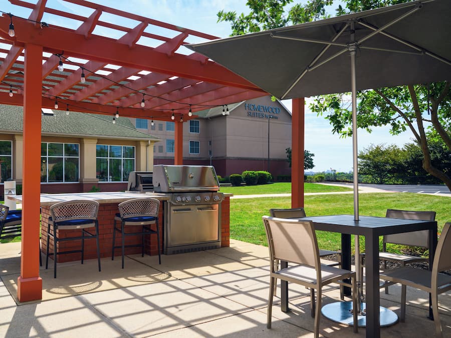 Een terras buiten met een barbecue, stoelen en een tafel onder een pergola.