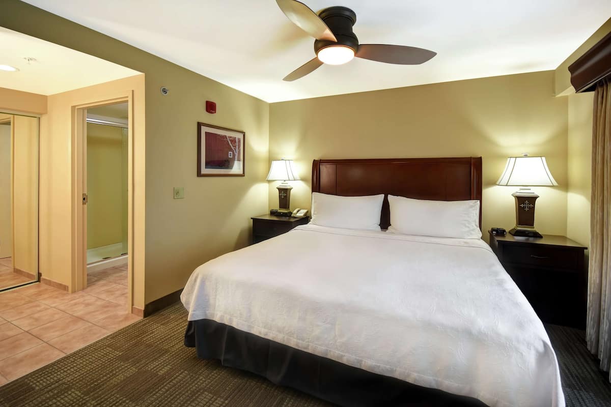 A hotel room with a large bed, two bedside tables with lamps, a ceiling fan, and a door leading to another room.