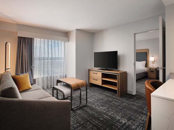 A hotel room with a sofa, a small table, a TV, a bed, a nightstand, and a mirror. - Homewood Suites Lancaster (Lancaster)