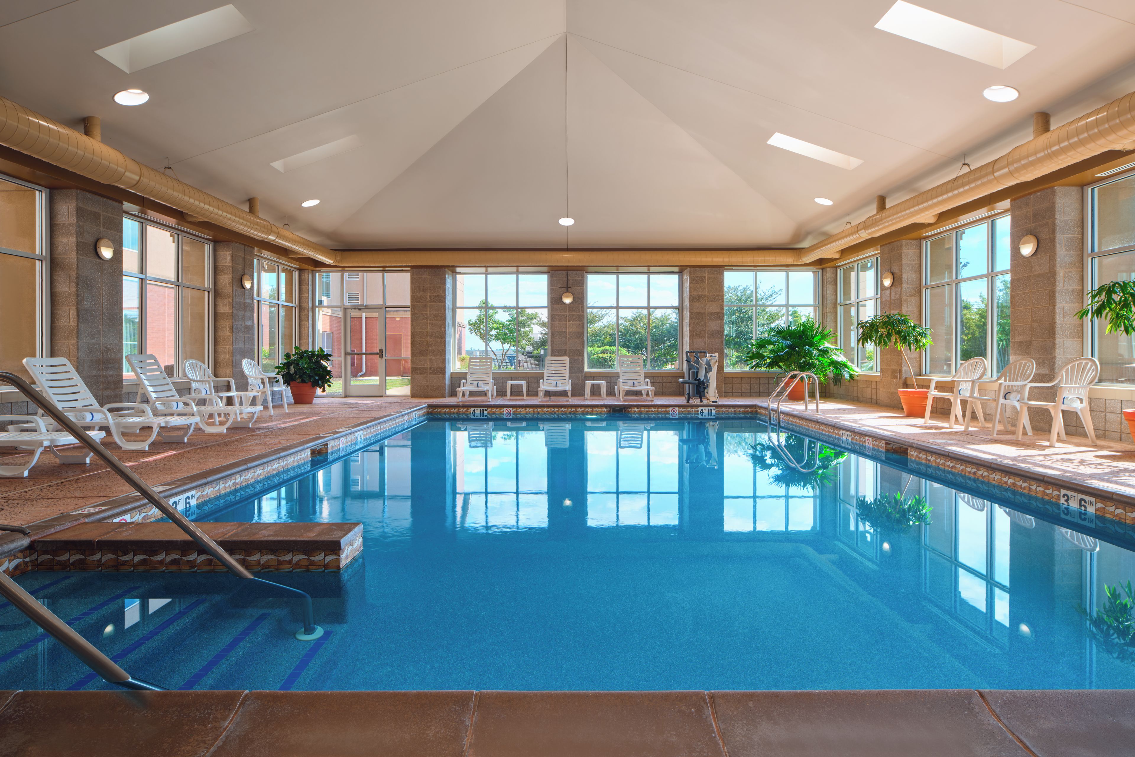 An indoor swimming pool with clear blue water, surrounded by white lounge chairs and potted plants.