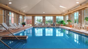 An indoor swimming pool with clear blue water, surrounded by white lounge chairs and potted plants.