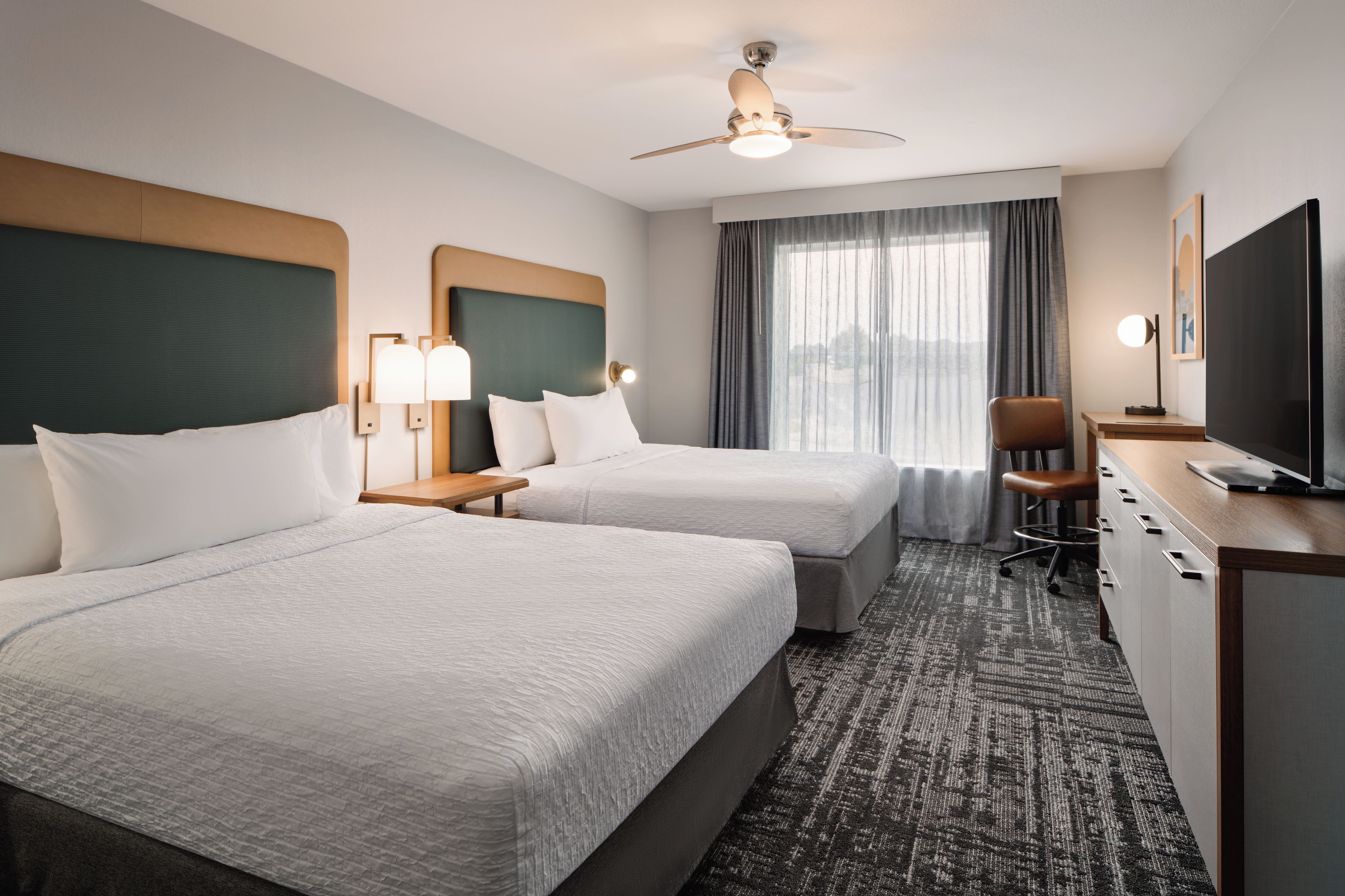 A hotel room with two beds, a desk, a chair, a television, and a ceiling fan.