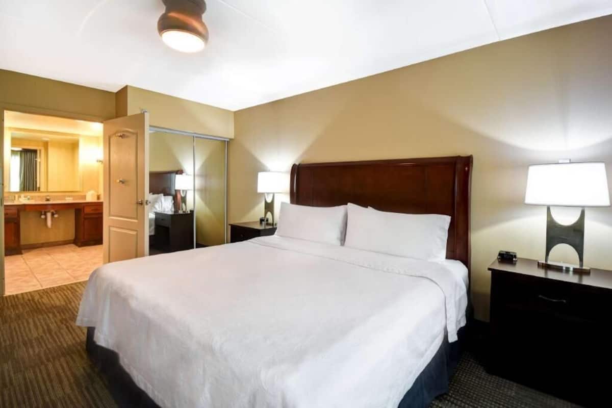 A hotel room with a large bed, a bedside table with a lamp, a nightstand, a mirror, and a bathroom visible through an open door.