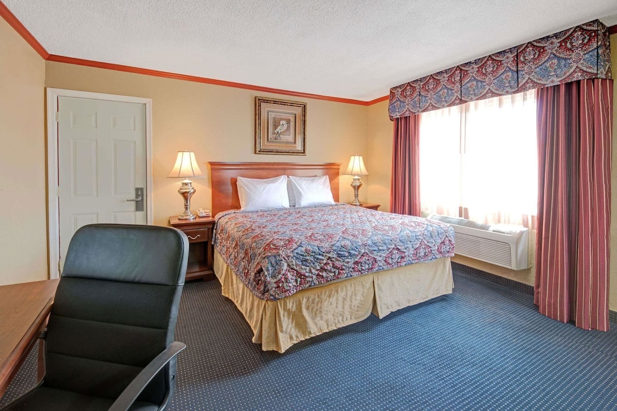 A hotel room with a bed, two bedside lamps, a chair, a door, and a window with curtains.