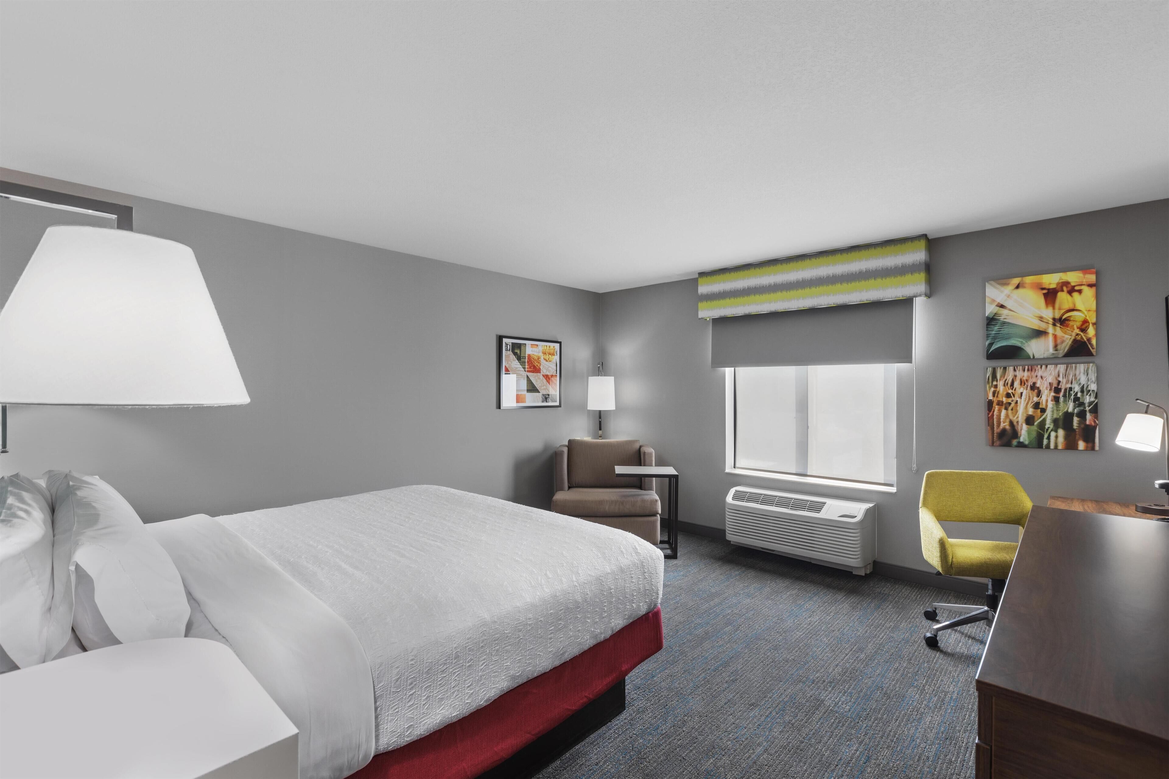 A hotel room with a bed, a desk, a chair, a lamp, a window, and artwork on the walls.