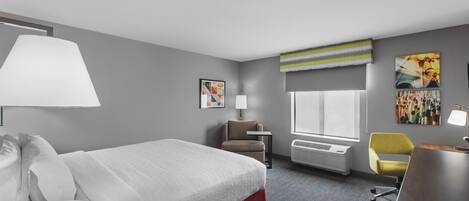 Habitación de hotel con cama, escritorio, silla, lámpara, ventana y obras de arte en las paredes.