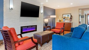 Un rebedor d'hotel modern amb un televisor de pantalla plana muntat a una paret amb relleu, una zona de seients amb un sofà blau i cadires taronja, una taula petita amb una làmpada, i una zona de bar al fons.