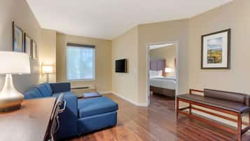 Una habitación de hotel moderna con piso de madera, un sofá azul, un banco de madera, un escritorio con lámpara y un televisor montado en la pared.