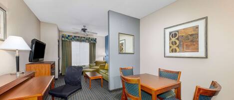 A hotel room with a wooden desk, a black office chair, a dining table with chairs, a television, and a ceiling fan.