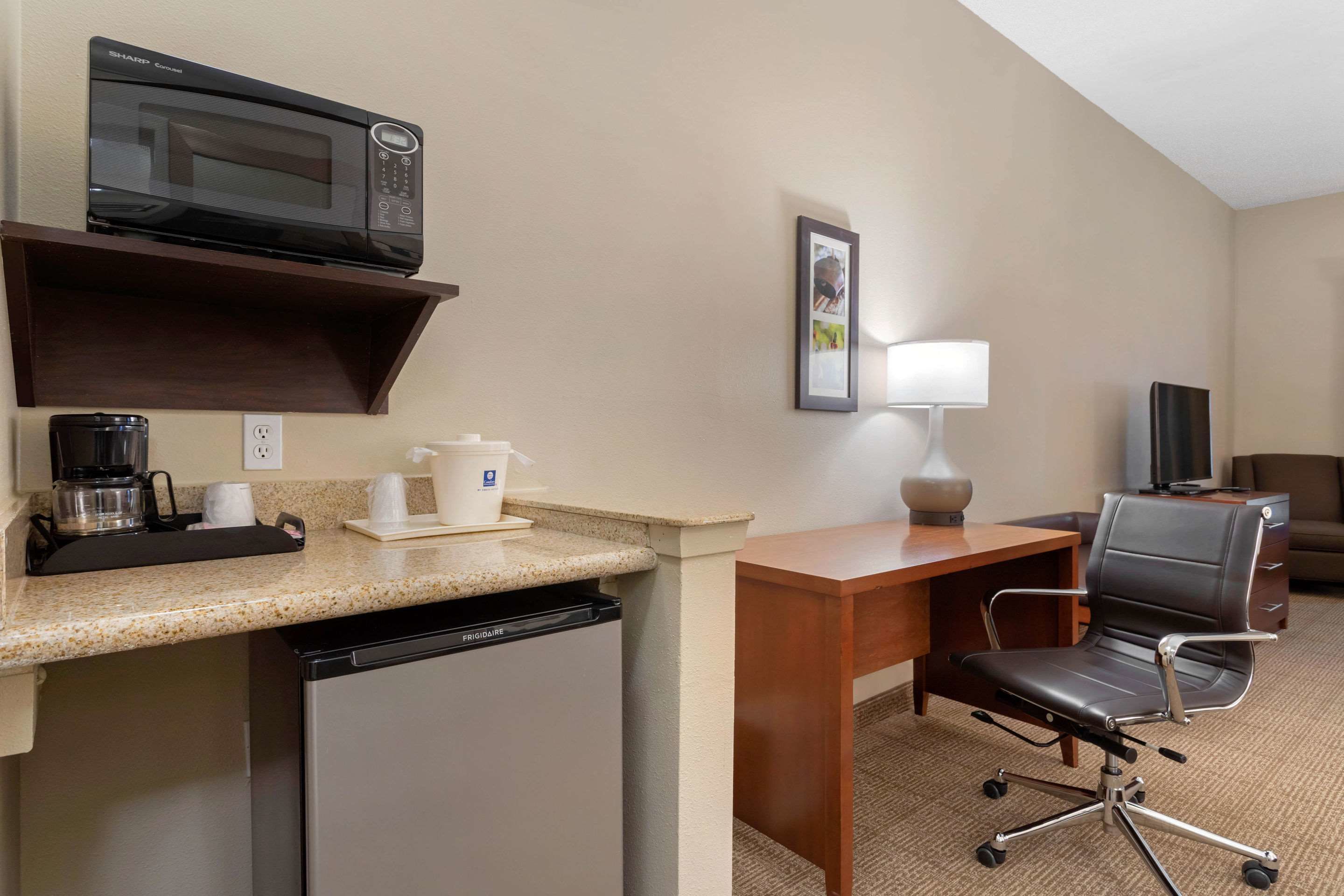 A compact hotel room with a kitchenette, including a microwave, coffee maker, and refrigerator. There is a desk with a chair and a television.