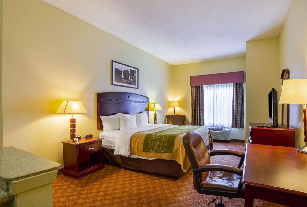 A hotel room with a bed, a desk, a chair, two bedside lamps, a television, and a window with curtains.