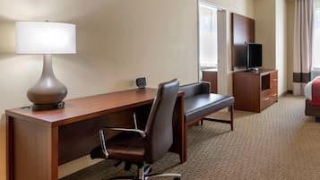 A hotel room with a wooden desk, a chair, a lamp, a television, a dresser, and a bed.